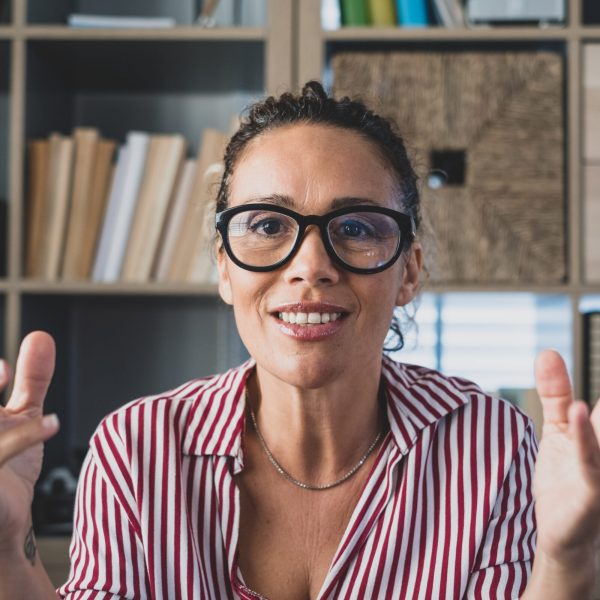 Head shot portrait confident businesswoman coach wearing glasses looking at camera and talking, mentor speaker holding online lesson, explaining, sitting at wooden work desk in modern cabinet
