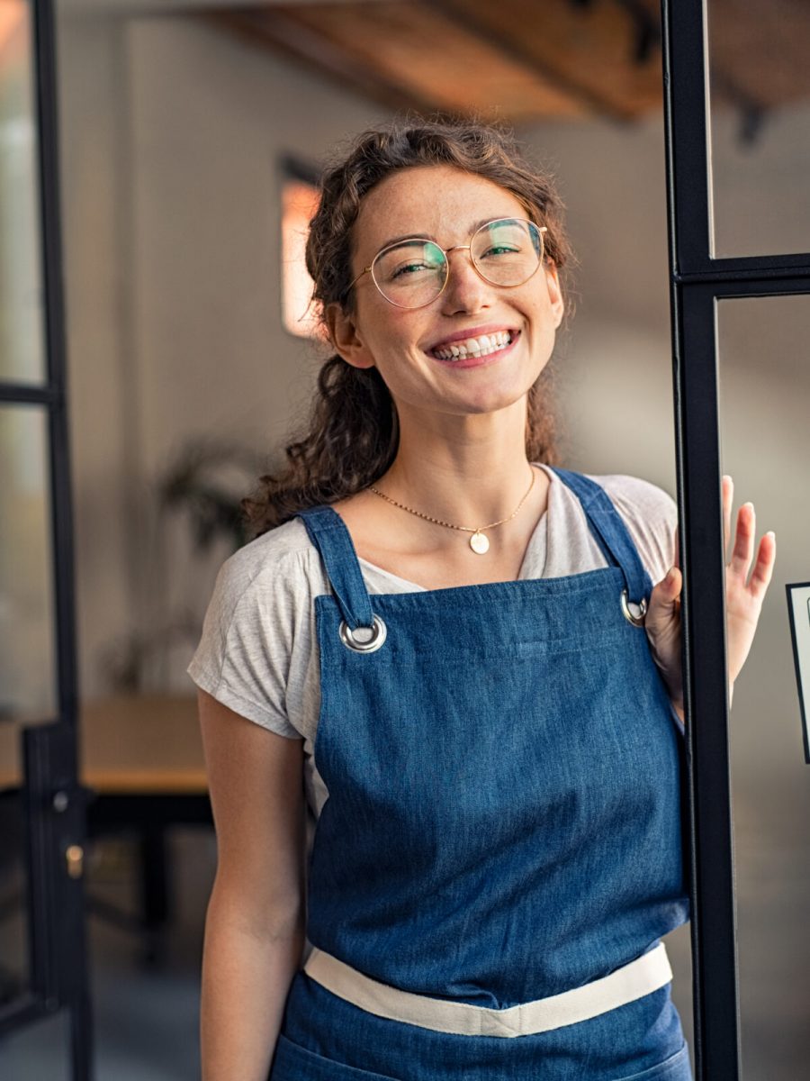 Portrait of positive business woman in apron standing at cafeteria door entrance. Cheerful young waitress in blue apron standing at glass door with open signboard and looking at camera. Excited small business owner standing at entrance of newly opened restaurant with open sign board.
